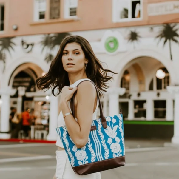 Tissli cerulean blue handwoven rug tote with white floral embroidery, brown leather base, and gold charm.