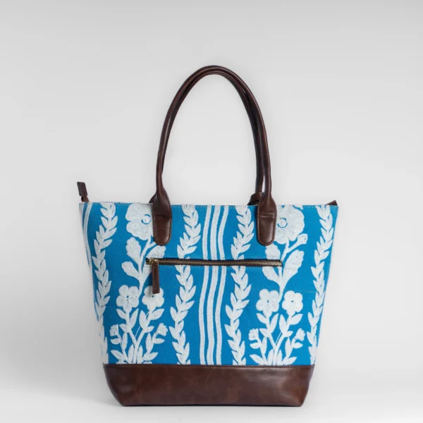 Tissli cerulean blue handwoven rug tote with white floral embroidery, brown leather base, and gold logo charm.