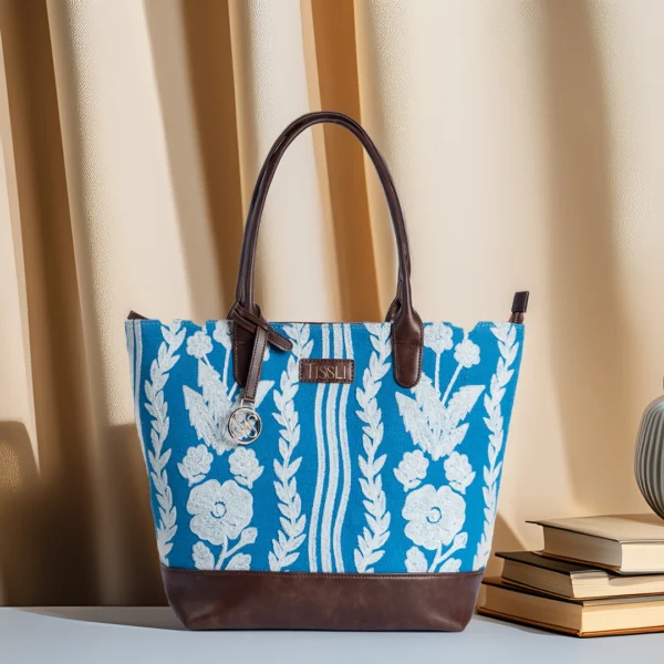 Tissli cerulean blue handwoven rug tote with white floral and vine embroidery, brown leather base, and gold logo charm.